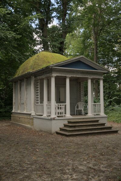 Fotografi af en hvid pavillion i neoklassisistisk stil, der står midt i en skov ved sommertid